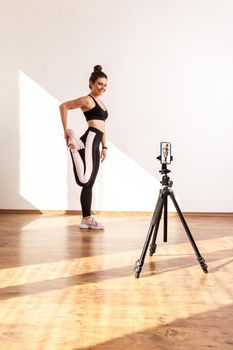Athletic coach standing on one leg, stretching muscles before training, recording video for her vlog, wearing black sports top and tights. Full length studio shot illuminated by sunlight from window.