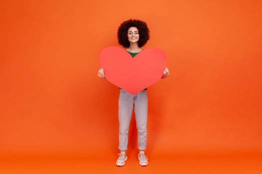 Full length portrait of falling in love woman with Afro hairstyle wearing green casual style sweater holding big red heart, looking with toothy smile. Indoor studio shot isolated on orange background.