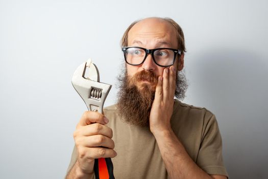 Male pull out tooth with wrench, looking at tool with confused expression, touching cheek with palm, bald bearded man wearing T-shirt and glasses. Indoor studio shot isolated on gray background.