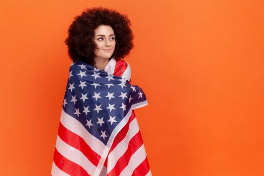 Calm dreamy woman with Afro hairstyle wearing green casual style sweater standing wrapped in USA flag, looking away, human rights. Indoor studio shot isolated on orange background.