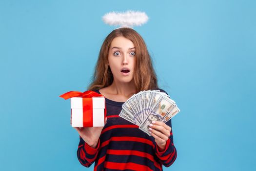 Astonished woman wearing striped casual style sweater and nimb over head, holding wrapped gift box and big fan of money, surprised by winning. Indoor studio shot isolated on blue background.