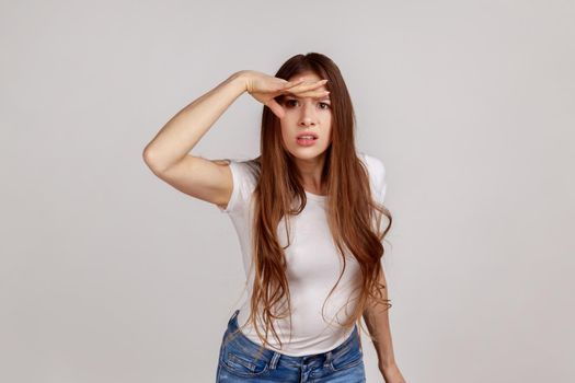 Portrait of concentrated attentive beautiful young woman standing seriously, with hand on forehead and looking far, wearing white T-shirt. Indoor studio shot isolated on gray background.