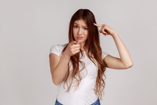 You're idiot with insane crazy plan. Displeased woman showing stupid gesture and pointing to camera, accusing of dumb idea, wearing white T-shirt. Indoor studio shot isolated on gray background.