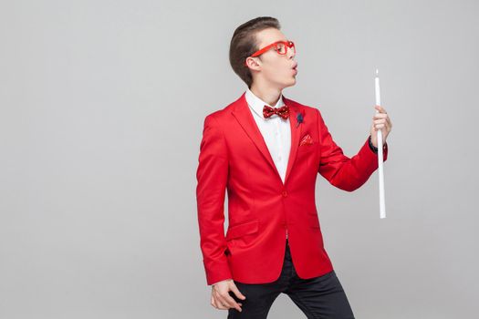 Portrait of elegant gentleman with trendy glasses and stylish hairdo in red tuxedo and bow tie blowing out big burning candle, romantic atmosphere. indoor studio shot isolated on gray background
