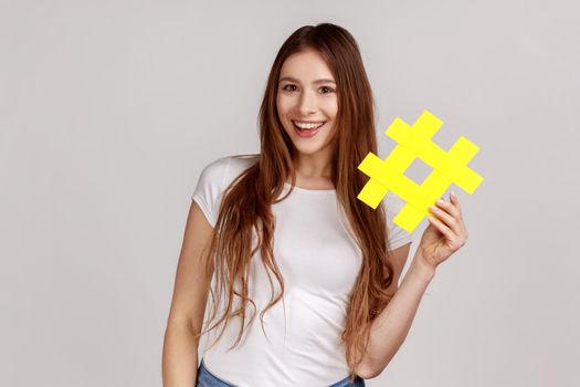 Smiling beautiful woman holding yellow hashtag symbol, making important topic popular, setting trends on Internet, wearing white T-shirt. Indoor studio shot isolated on gray background.