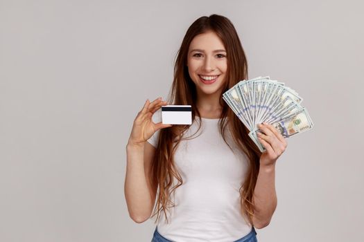 Portrait of smiling woman holding credit card and big fan of dollar banknotes, banking, earning money, wearing white casual style T-shirt. Indoor studio shot isolated on gray background.