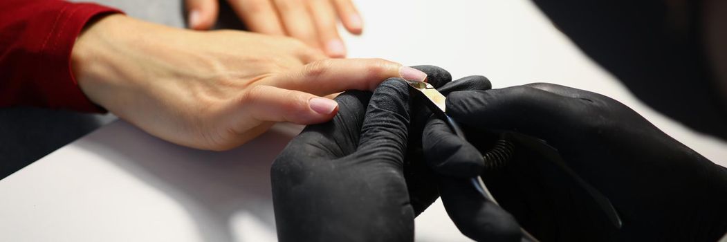 Close-up of nail master cutting off cuticle on clients hands with special equipment. Professional work in black gloves. Beauty industry, wellness concept