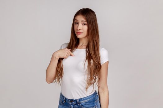 This is me. Portrait of arrogant woman pointing herself and looking selfish egoistic haughty, feeling proud of own achievement, wearing white T-shirt. Indoor studio shot isolated on gray background.