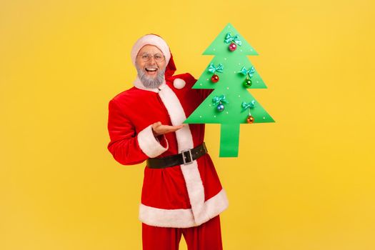 Happy elderly man with gray beard wearing santa claus costume standing holding green paper eco tree, looking at camera with happy smile. Indoor studio shot isolated on yellow background.