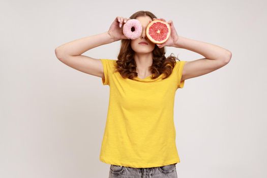 Sugary dessert vs fruits. Portrait of playful brown haired woman in yellow T-shirt covering eyes with fresh grapefruit and sweet doughnut, junk food. Indoor studio shot isolated on gray background.