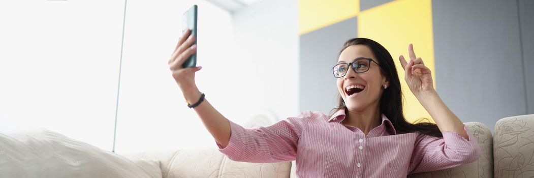 Low angle of pretty young female show peace sign and take selfie on smartphone. Talk to friend or family on video call. Technology, fun, leisure concept