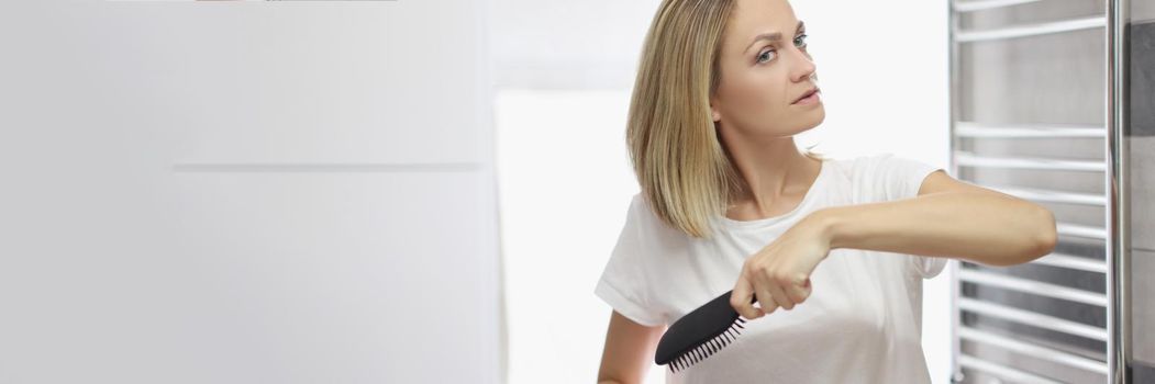 Portrait of young blonde woman looking in mirror and brushing hair in bathroom, no makeup, morning routine, natural beauty. Good morning, wellness concept