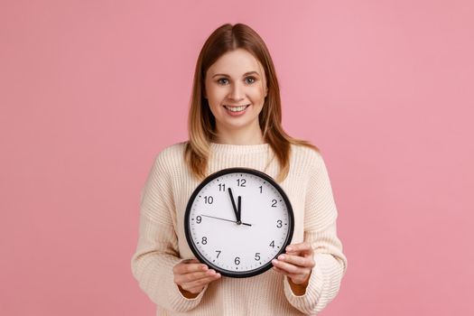 Portrait of happy positive blond woman holding big wall clock, looking at camera with pleasant smile, time to go, wearing white sweater. Indoor studio shot isolated on pink background.