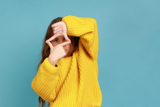 Portrait of little girl looking through photo frame hand gesture, focusing picture, observing world, wearing yellow casual style sweater. Indoor studio shot isolated on blue background.