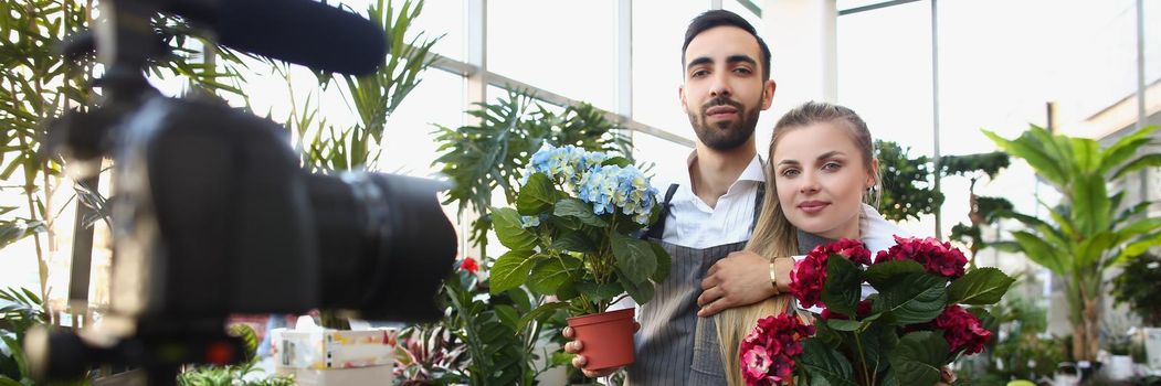 Portrait of couple record video for social media content, beautiful photoshot in botanical garden with flowers. Man and woman hold plants. Modeling concept