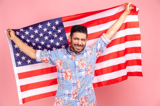 Smiling man with beard wearing casual style blue shirt, holding USA flag and looking at camera with rejoicing look, celebrating national holiday. Indoor studio shot isolated on pink background.