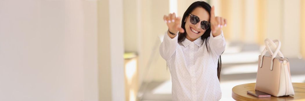 Portrait of cheerful happy young woman in stylish outfit show some symbols with hands. Attractive lady sit outdoors, gesture on camera. Trendy, joy concept