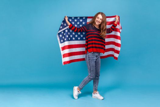 Full length portrait of happy woman in striped casual style sweater, holding USA flag and looking at camera with smile., celebrating national holidays. Indoor studio shot isolated on blue background.