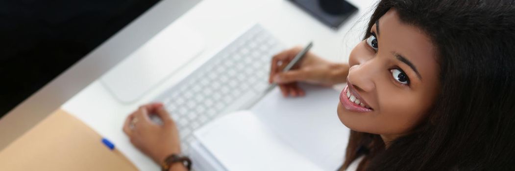 Top view of smiling afro american young woman on workplace, open journal, luxury pc device on desk. Fresh start, mockup, copy space, technology concept