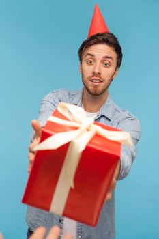 POV hand giving wrapped red gift box to bearded man with party cone on head, guy wearing denim shirt looking at birthday present surprise with delight. Indoor studio shot isolated on blue background.