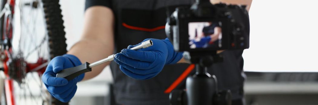 Close-up of blogger mechanic fix bicycle in workshop, busy young worker in shirt and gloves show modern cycle transmission system with spanner on camera