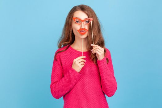 Portrait of positive woman in pink pullover, holding paper glasses and lips, looking at camera, having festive mood, wearing party props. Indoor studio shot isolated on blue background.
