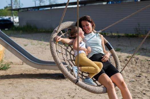 Mom and daughter swing on a round swing. Caucasian woman and little girl have fun on the playground