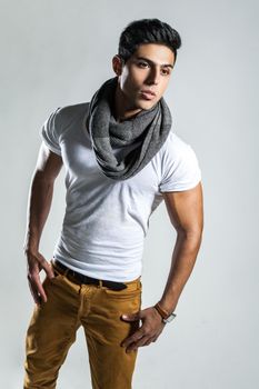 Portrait of handsome brunette sexy man wearing fashionable white T-shirt, scarf and trousers, looking away with confident expression. Indoor studio shot isolated on gray background.