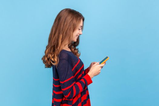 Side view of smiling optimistic woman standing with smart phone in hands, answering romantic message from boyfriend, wearing striped casual sweater, Indoor studio shot isolated on blue background