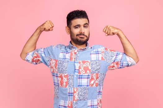 I'm strong. Portrait of attractive man in casual blue shirt raising hands to show biceps and looking confident at camera, feeling power and strength. Indoor studio shot isolated on pink background.