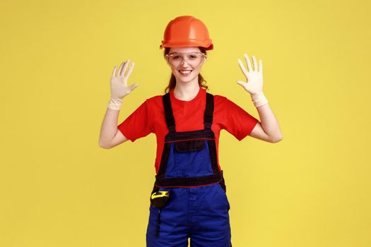 Positive smiling builder woman standing with raised arms, showing her rubber gloves for hands protecting, wearing overalls and protective helmet. Indoor studio shot isolated on yellow background.