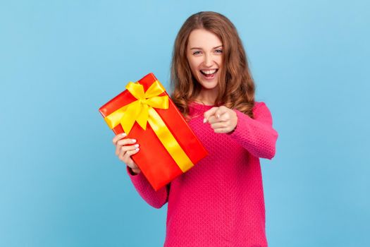 Hey, you are winner. Happy optimistic woman wearing pink pullover, holding present box and pointing finger to camera with excited expression. Indoor studio shot isolated on blue background.