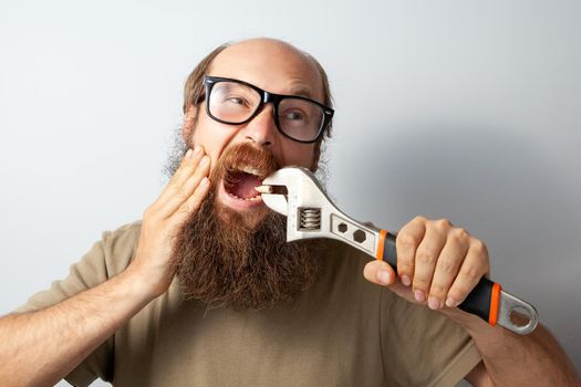 Adult male pulling out bad tooth by his own, touching cheek with hand, screaming from terrible pain, bald bearded man wearing T-shirt and glasses. Indoor studio shot isolated on gray background.