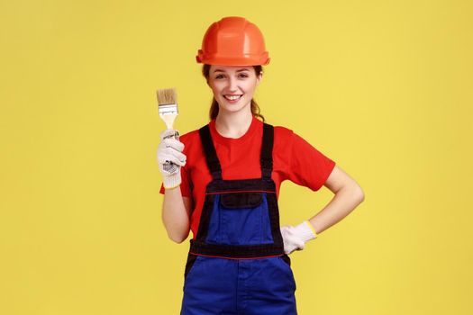 Positive young adult builder woman standing with paintbrush in hands, wearing work uniform and protective helmet, keeps hand on waist. Indoor studio shot isolated on yellow background.