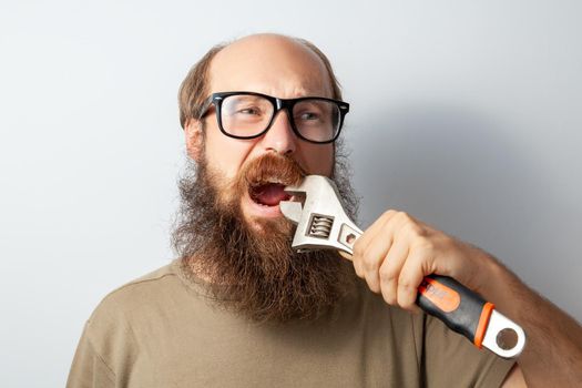 Portrait of male trying to pull out bad tooth with adjustable wrench , keeps mouth opened, bald bearded man wearing T-shirt and glasses. Indoor studio shot isolated on gray background.