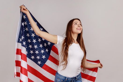 Portrait of happy positive woman with dark hair, holding american flag, looking away, celebrating national holiday, wearing white T-shirt. Indoor studio shot isolated on gray background.