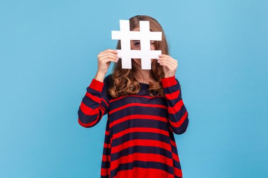 Curious woman wearing striped casual style sweater, looking at camera through large white hashtag symbol with prying eye, interesting web content. Indoor studio shot isolated on blue background.