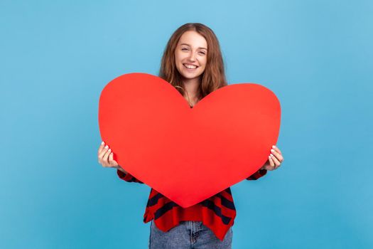 Happy woman wearing striped casual style sweater, holding big red heart and looking at camera with smile, falling in love, romantic feelings. Indoor studio shot isolated on blue background.