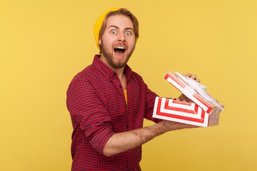 Portrait of hipster bearded guy in beanie hat and checkered shirt holding opened gift box and looking at camera with amazed smile, unpacking present. Indoor studio shot isolated on yellow background.