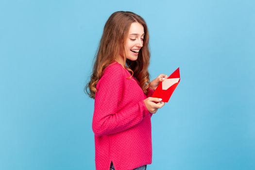 Side view of happy woman wearing pink pullover, reading letter or greeting card, holding envelope, smiling and rejoicing pleasant news. Indoor studio shot isolated on blue background.