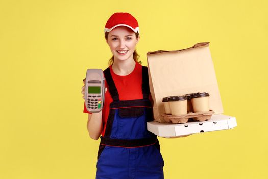 Portrait of optimistic courier woman holding terminal and coffee with pizza box, looking at camera with happy expression, wearing overalls and cap. Indoor studio shot isolated on yellow background.