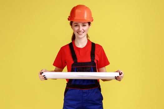 Portrait of young ambitious woman architect holding rolled blueprints in hands, project of new building, wearing work uniform and protective helmet. Indoor studio shot isolated on yellow background.