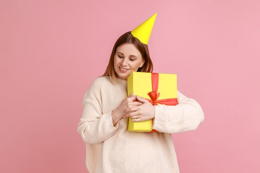 Portrait of happy cute blond woman in party cone embracing yellow gift box, getting present from friend on her birthday, wearing white sweater. Indoor studio shot isolated on pink background.