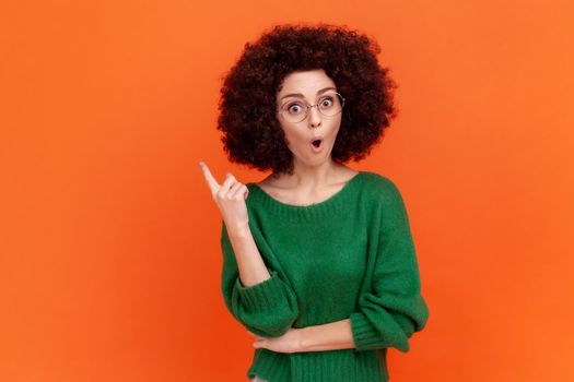 Excited woman with Afro hairstyle wearing green casual style sweater and glasses standing with open mouth and raised finger, having idea. Indoor studio shot isolated on orange background.