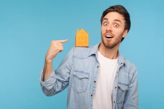 Portrait of amazed astonished man wearing denim shirt, standing pointing at paper house on his shoulder and smiling at camera, home purchase. Indoor studio shot isolated on blue background.