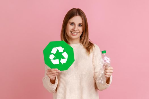 Happy optimistic young adult blond woman holding green recycling sign and empty plastic bottle, calls on to protect environment, wearing white sweater. Indoor studio shot isolated on pink background.