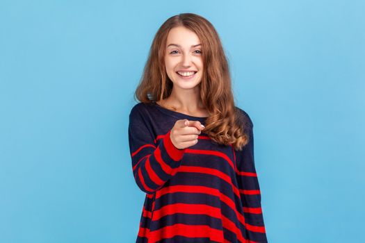 Hey you. Happy woman wearing striped casual style sweater pointing to camera and looking with charming toothy smile, indicating finger, making choice. Indoor studio shot isolated on blue background.
