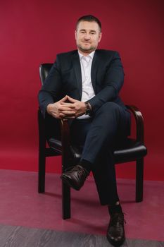Portrait of a business man in a black suit sitting in a studio on a chair against an empty wall.