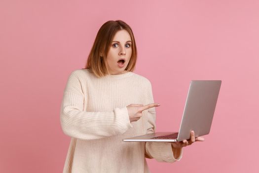 Portrait of astonished shocked young adult blond woman working on laptop compute, looking at camera and pointing at display, wearing white sweater. Indoor studio shot isolated on pink background.