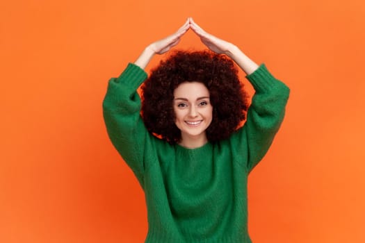 Woman with Afro hairstyle wearing green casual style sweater making house roof gesture and smiling, concept of life insurance, protection. Indoor studio shot isolated on orange background.